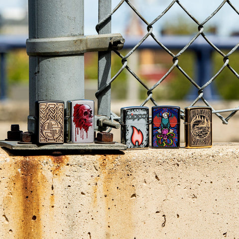 Lifestyle image of Bloody Skull Design Brushed Chrome Windproof Lighter standing with four other lighters in front of a chain link fence