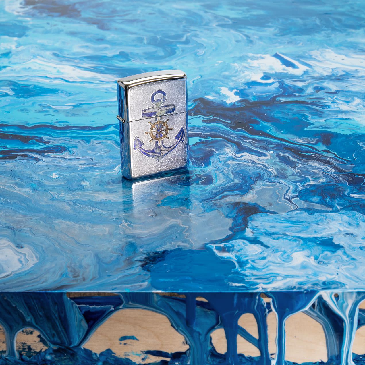 Lifestyle image of Anchor Design High Polish Chrome Windproof Lighter standing on a blue piece of art